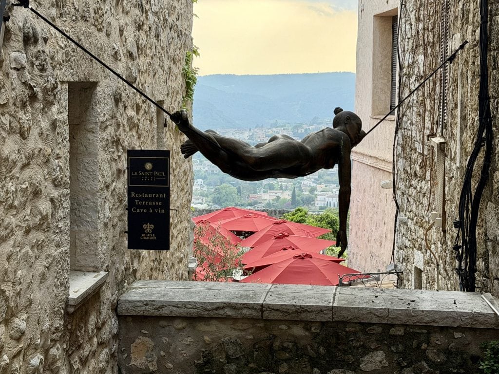 A metal sculpture of a naked woman, fully relaxed, laying face down, suspended precariously on a rope between two buildings.